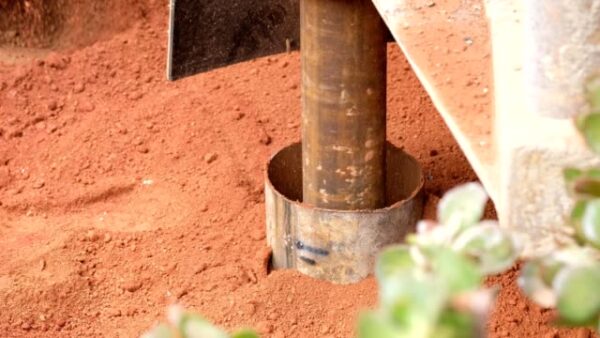 A large borehole drilling machine works through the loose clay in an effort to sink a borehole, large amounts of red clay soil spill out of the hole.